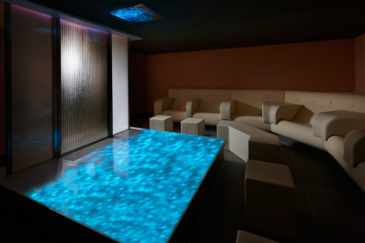 Glowing blue floor panel emits wave-like light, illuminating a dim lounge. Cream sectional sofas and cube stools surround it; frosted sliding panels and a ceiling light complete a modern room.